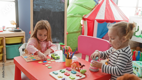 Cuadro en lienzo Two little girls placing wooden shape pieces and stacking rings on a red table, sorting toys and pointing in a children's playroom building; concentration friendship