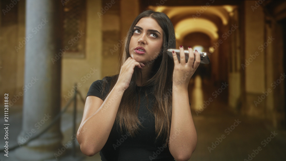 Obraz premium Young woman holding smartphone to ear with hand on chin in building corridor, looking upward and thoughtful; contemplative.