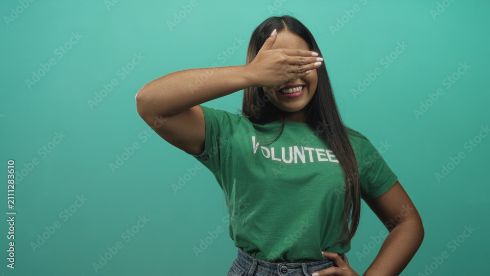 Fototapeta premium Young hispanic woman wearing green volunteer tshirt covering eyes with hand while smiling and hand on hip in studio; community service joy.