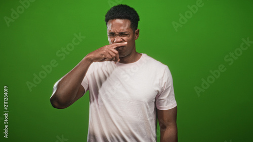 Quadro su tela Man pinches nose in studio with green backdrop and pink tshirt, grimacing at a foul smell; disgust discomfort