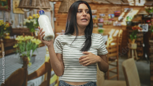 Foto Young hispanic woman holds milk bottle aloft with hand on hip and pensive gaze, wearing striped top in a rustic cafe dining area; skepticism consumer choice