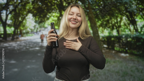 Quadro su tela Woman holding beer bottle, smiling with eyes closed, right hand on chest on forest path beside trees and hedge; gratitude contentment
