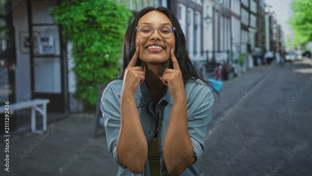 Fototapeta premium Woman points index fingers to cheeks while smiling, wearing yellow top and denim shirt on a street; joy playfulness.