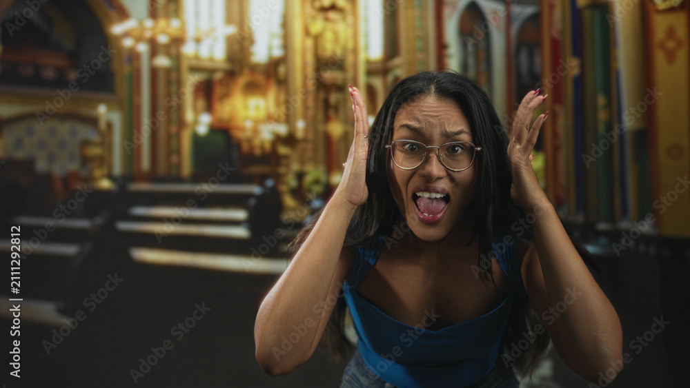 Fototapeta premium Young hispanic woman in blue top wearing glasses pressing hands to head and sticking out tongue in ornate church building; surprise playfulness.