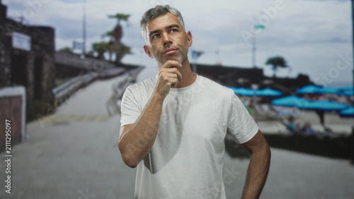 Fotografie Man hand on chin pensive pose on beach promenade with blue umbrellas and walkway; thoughtful reflection