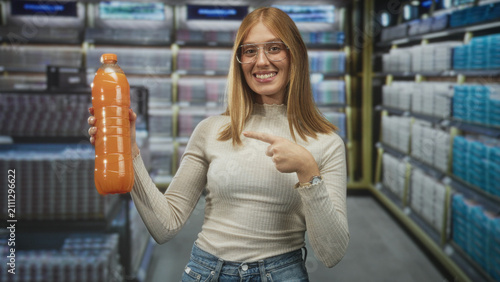 Photography Redhead woman pointing finger at orange juice bottle in supermarket aisle, holding the plastic bottle near stocked shelves of drinks while smiling and wearing glasses and jeans; cheerful endorsement