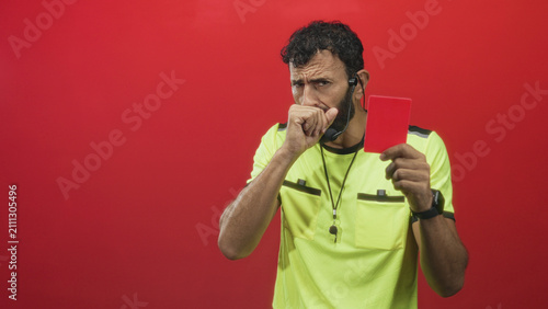 Obraz na płótnie Man referee in neon yellow shirt holds red card and coughs into his fist in studio, wearing whistle on lanyard and earpiece with serious expression; concern
