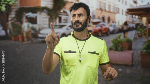 Bild auf Leinwand Man referee in neon yellow shirt pointing index finger with whistle on necklace and earpiece, beard visible in street; authority control focus