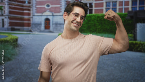 Fotografía Man flexes bicep and smiles while raising fist in front of a redbrick building entrance, wearing glasses and short sleeve shirt; confidence casual