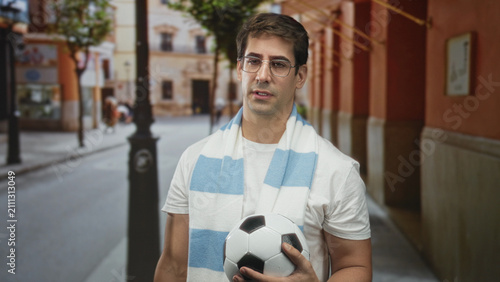 Canvas Print Man holding soccer ball with blue and white striped scarf, standing on a narrow street near archways and a lamppost; anticipation fandom