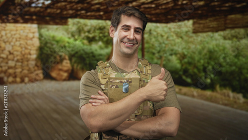 фотографія Man soldier wearing camouflage vest and dogtags, arms crossed beneath a camo canopy on a forest deck; pride duty