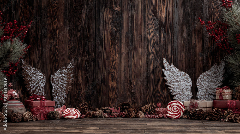 Obraz premium Festive Christmas background on dark wood featuring pine branches, cones, candy canes, red berries, angel wings, and gifts at top with copy space. 