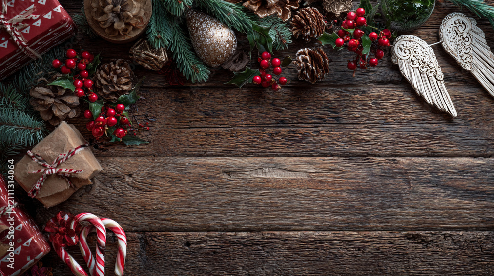 Obraz premium Festive Christmas background on dark wood featuring pine branches, cones, candy canes, red berries, angel wings, and gifts at top with copy space. 