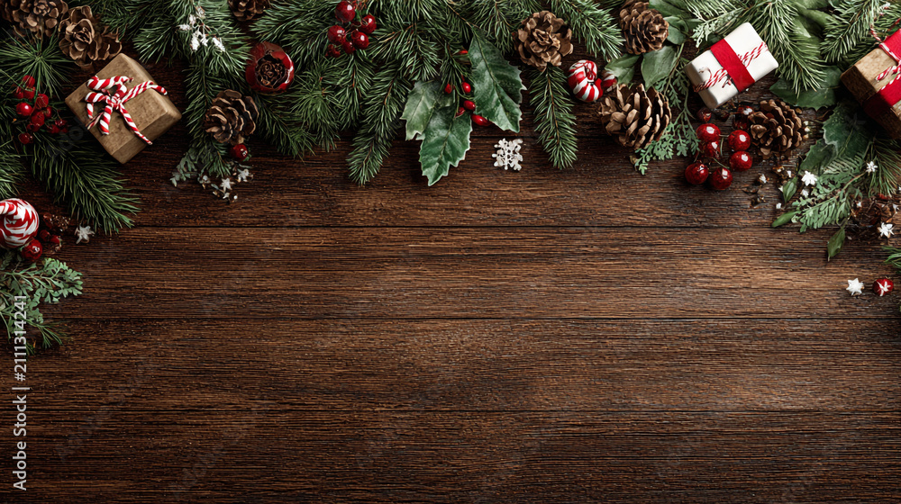Obraz premium Festive Christmas background on dark wood featuring pine branches, cones, candy canes, red berries, angel wings, and gifts at top with copy space. 