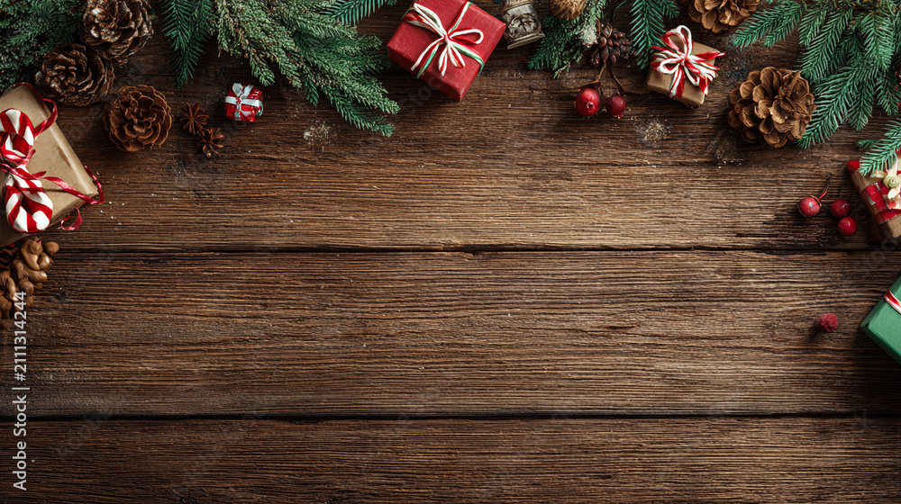 Obraz premium Festive Christmas background on dark wood featuring pine branches, cones, candy canes, red berries, angel wings, and gifts at top with copy space. 