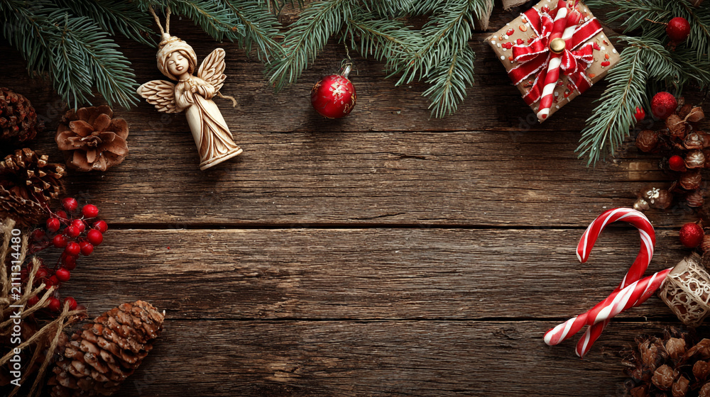 Obraz premium Festive Christmas background on dark wood featuring pine branches, cones, candy canes, red berries, angel wings, and gifts at top with copy space. 