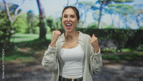 Bild auf Leinwand Woman in white tanktop and striped overshirt smiling with fists raised and pointing to her temple, casual jeans and belt, standing on a sunlit park path in a forest; joy