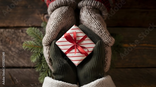 Top view of hands in warm knitted mittens holding a small gift box wrapped in striped paper with a red ribbon on a rustic wooden background with pine branches