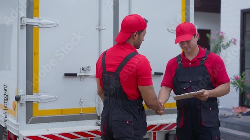 Delivery worker in red uniform carrying cardboard box to cargo truck at loading dock while logistics team discusses shipment service with clipboard beside vehicle