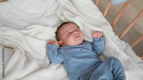 фотографія Baby boy sleeping with clenched fists and curled legs in a blue knit outfit on a white fleece blanket inside a wooden crib; gentle serenity