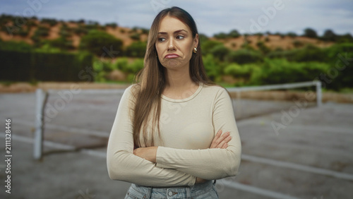 Quadro su tela Woman with crossed arms and pouting lips standing on an outdoor tennis court near a net and posts; sullen disappointment