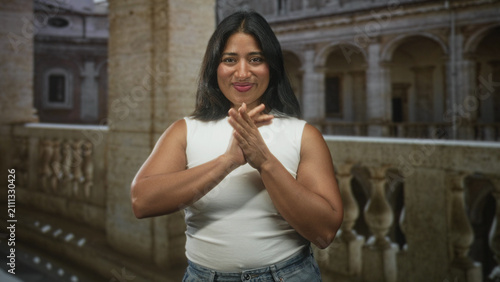 Фотография Woman presses hands together in prayer with visible hands and bare arms, smiling on a stone balustrade of a historic building; gratitude
