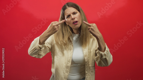 Foto Woman displays raised middle fingers and a stern facial expression toward camera in a red studio backdrop, wearing a beige shirt and white top; defiance rebellion