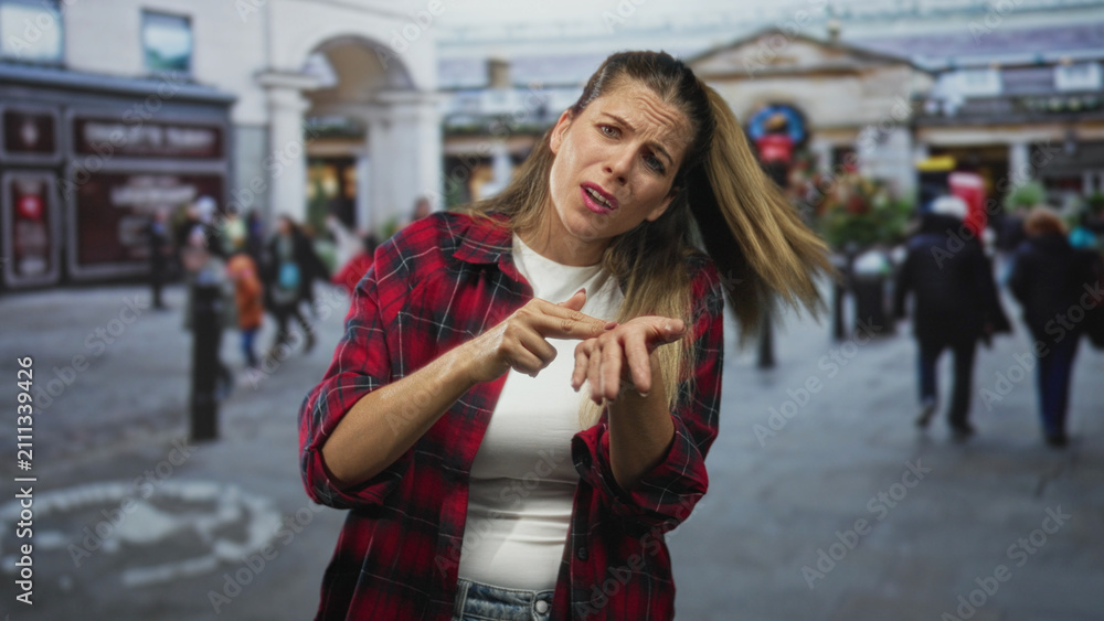 Obraz premium Woman in a red plaid shirt holds hand out with pinched fingers and palm up, asking gesture in a busy street plaza; frustration plea.