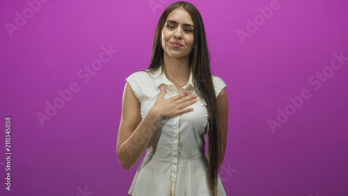 Young woman with hand on chest touching her palm in studio under purple lighting and slight smile; gratitude sincerity.
