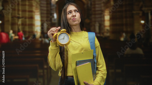 фотографія Young female student in yellow sweater holds a yellow alarm clock and folders with a blue backpack, pouting while raising the clock in a building with pews; lateness anxiety
