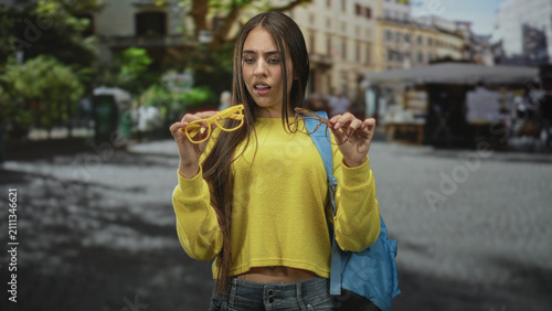 Canvastavla Young woman holds two pairs of eyeglasses while standing in a cobblestone street market, yellow sweater and blue backpack visible; surprise indecision