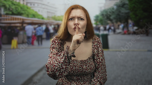 Konstfotografi Redhead woman pressing index finger to lips for silence while standing in a crowded urban street market with blurred pedestrians behind her; warning secrecy