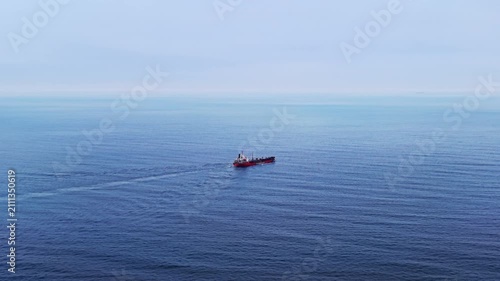 Red cargo ship crossing calm sea. Wide horizon composition with soft blue water and a faint wake trailing behind the vessel.