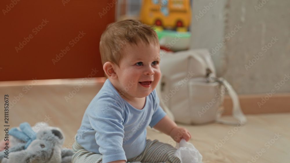 Fototapeta premium Boy holding wooden toy and white cloth with hand while sitting on floor in building near plush rabbit and diaper bag; joyful play.