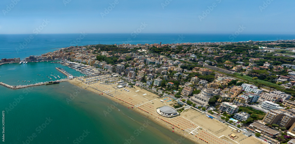 Fototapeta premium Aerial view of Anzio, in Lazio, Italy. It's a coastal city featuring a busy harbor, sandy beaches, and urban buildings along the shore under a clear sky, capturing an ideal summer destination.