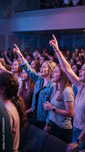 A group of diverse young people singing and raising their hands with joy at a modern indoor church service or youth conference.