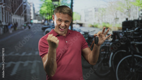 Obraz na plátne Man in red polo holds car keys aloft with a clenched fist, grinning while pumping his arm on a street lined with parked bicycles and canal buildings; triumph success