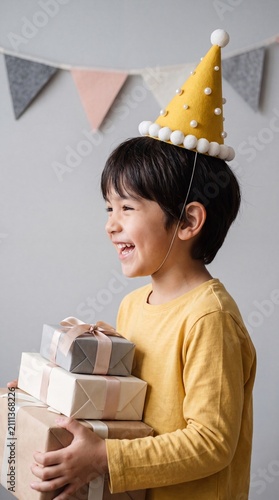A cheerful young Asian boy wearing a festive yellow party hat holds a stack of wrapped birthday gifts while smiling, against a neutral background.