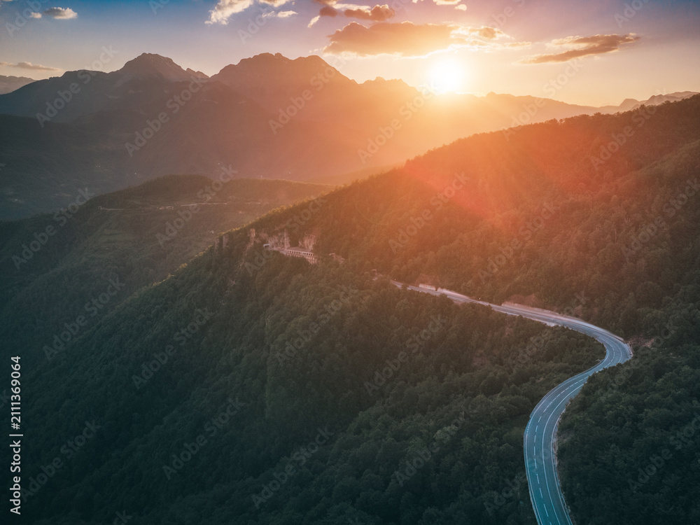 Obraz premium Winding mountain road traversing a lush green forest with dramatic sun flare and mountain peaks during a vibrant sunset