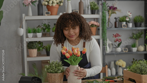 Картината върху платно Woman florist arranges orange tulips in a ceramic vase in a studio, hands adjusting stems and placing bouquet with care; calm focus