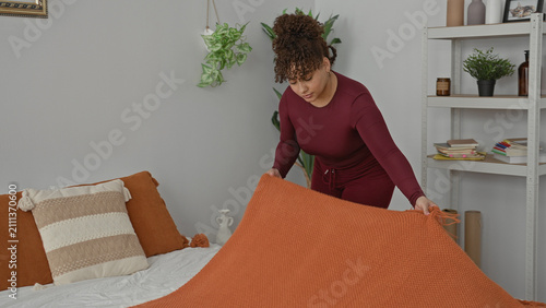 Fotografie Woman smoothing orange blanket and straightening pillows on bed inside building; calm domestic routine