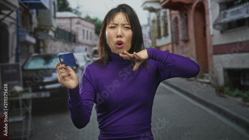 Woman holding creditcard with raised hand shielding face on a narrow street, puckered lips and dismissive gesture; rejection caution.