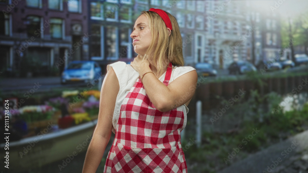 Fototapeta premium Woman in red gingham apron holding neck with hand showing neck pain on a street; quiet persistent discomfort.