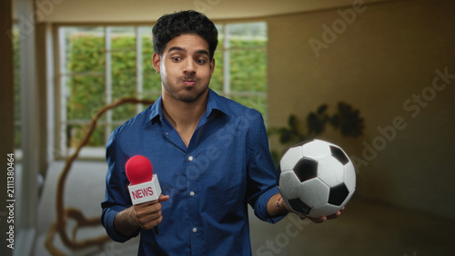 Fotografie Man holding news microphone and soccer ball, looks down with a slight smile in studio; playful curiosity