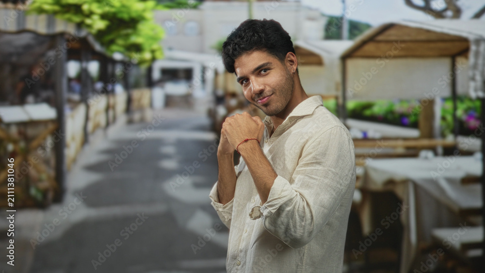Fototapeta premium Man in a light linen shirt smiles with clasped hands near chest at a busy street cafe market row, facing camera; joyful relaxed confidence.
