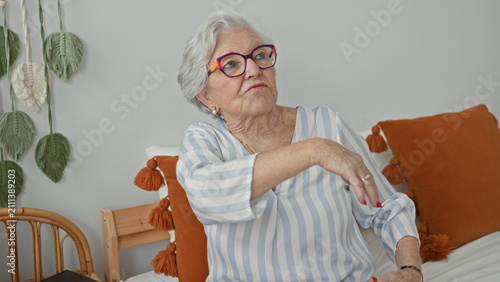 Obraz na płótnie Elderly woman wearing colorful glasses and striped blouse sits on bed among orange cushions, waves blurred hand across face in studio; gentle nostalgia