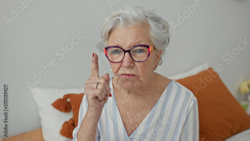 Foto Elderly woman points finger while seated on bed in a bedroom building, wearing colorful glasses and pearl earring; disapproval