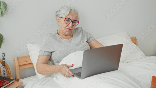 Foto Senior woman with grey hair and glasses points finger to laptop screen while seated propped under duvet in bed inside building; focused comfort