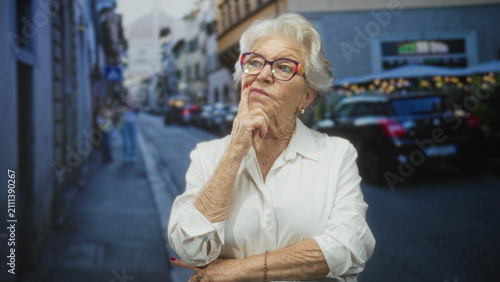 фотографія Old woman with colorful glasses touches chin with finger in a pensive thinking gesture on a narrow city street; thoughtful reflection