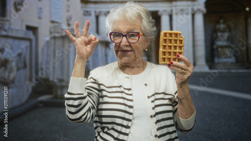 Quadro su tela Senior woman holding waffle and making ok hand gesture while smiling, wearing striped cardigan and colorful glasses in front of stone building; cheerful nostalgia